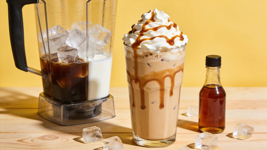 A caramel frappe topped with whipped cream and caramel drizzle beside a blender of coffee, milk and ice