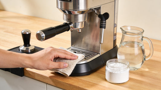 A hand wiping down a home espresso machine beside a jug of water and a jar of descaling powder