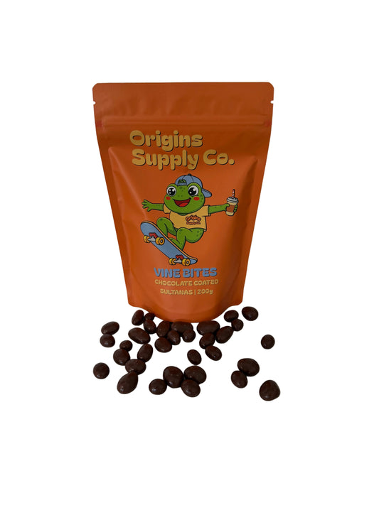 An orange stand-up pouch of Origins Supply Co. Vine Bites Chocolate Covered sultanas stands on a white marble countertop with several chocolate-coated banana pieces scattered in front.