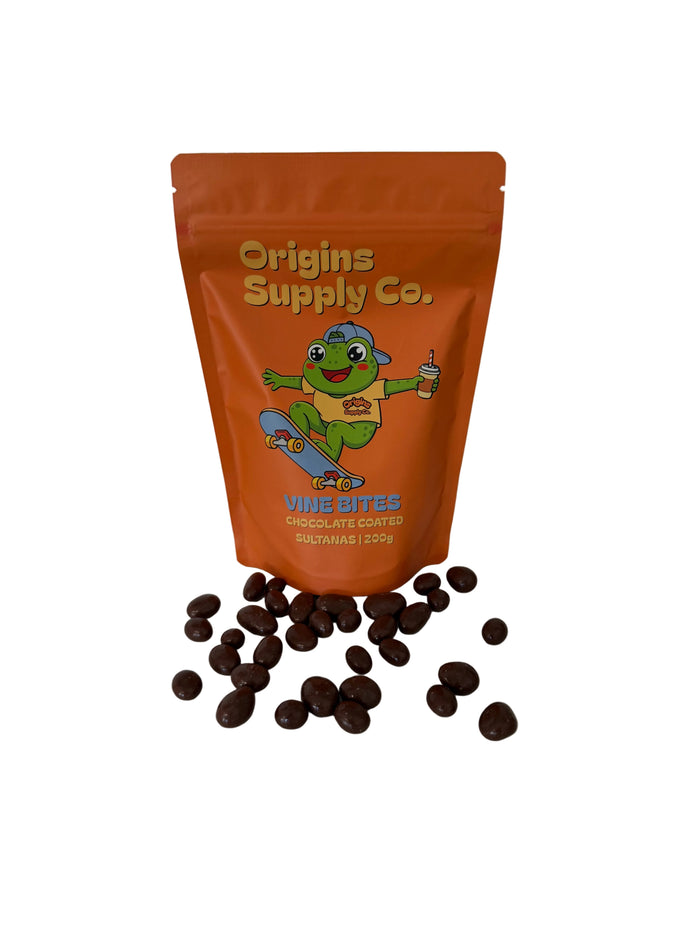 An orange stand-up pouch of Origins Supply Co. Vine Bites Chocolate Covered sultanas stands on a white marble countertop with several chocolate-coated banana pieces scattered in front.