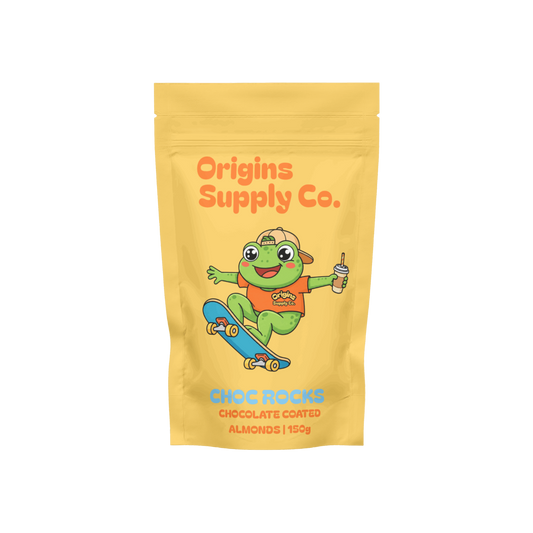 A yellow stand-up pouch of Origins Supply Co. Choc Rocks chocolate coated almonds, featuring a cartoon green frog character on a skateboard with an orange shirt and a blue skateboard.