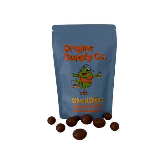 A blue stand-up pouch labeled 'Origins Supply Co. Weird Bites Chocolate Coq Au Lait' stands on a white marble countertop with several chocolate-coated bites scattered in front.