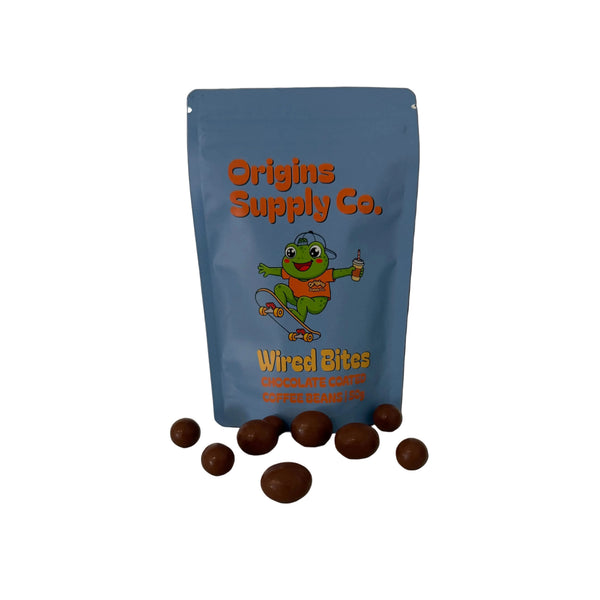 A blue stand-up pouch labeled 'Origins Supply Co. Weird Bites Chocolate Coq Au Lait' stands on a white marble countertop with several chocolate-coated bites scattered in front.